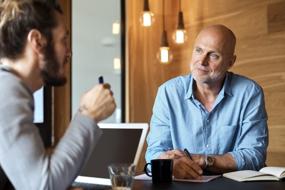 To menn som sitter på kontor og snakker sammen over en PC