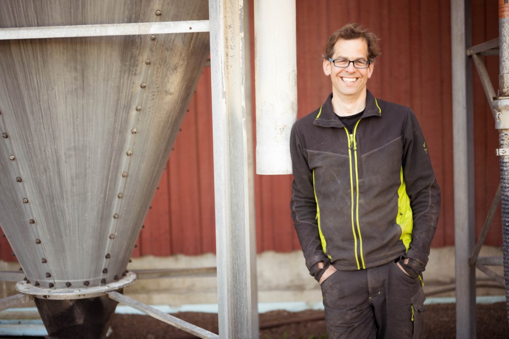 En glad bonde i en sort og limegrønn jakke står smilende foran en stor metallisk traktformet beholder, innenfor en driftsbygning. Bakgrunnen viser den røde veggen til bygningen, og understreker et typisk landbruksscenario.