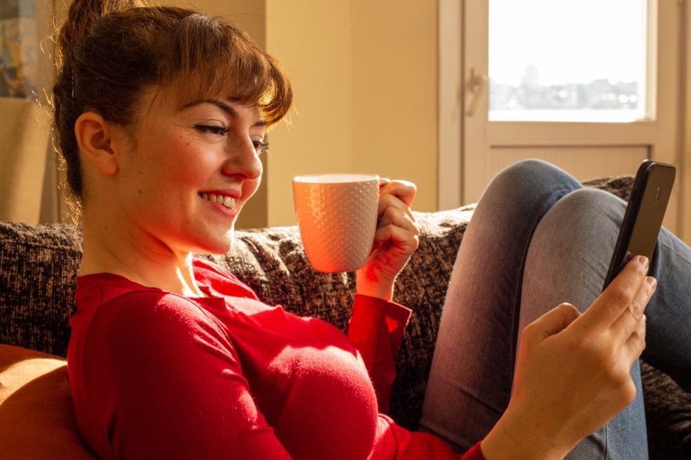 Kvinne i rødt sitter i sofaen og smiler mens hun ser på mobilen og holder en kaffekopp i vårsol. Illustrerer kontroll og ro i hverdagsøkonomien.