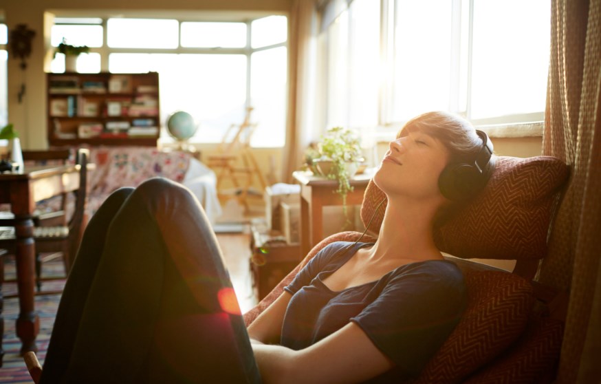 Ung kvinne sitter i lenestol i stue og lytter til headset